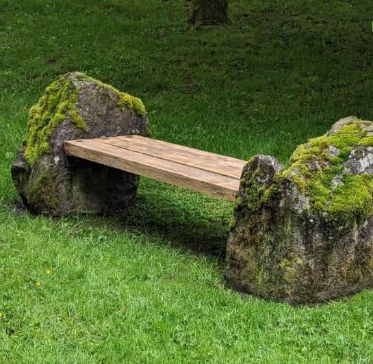 An outdoor bench composed of three wooden planks forming the seat, supported by two large, moss-cove… — 3D model