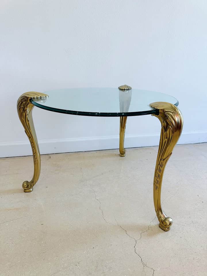 A round accent table featuring a clear glass top supported by three ornate, gilded metal legs design… — 3D model