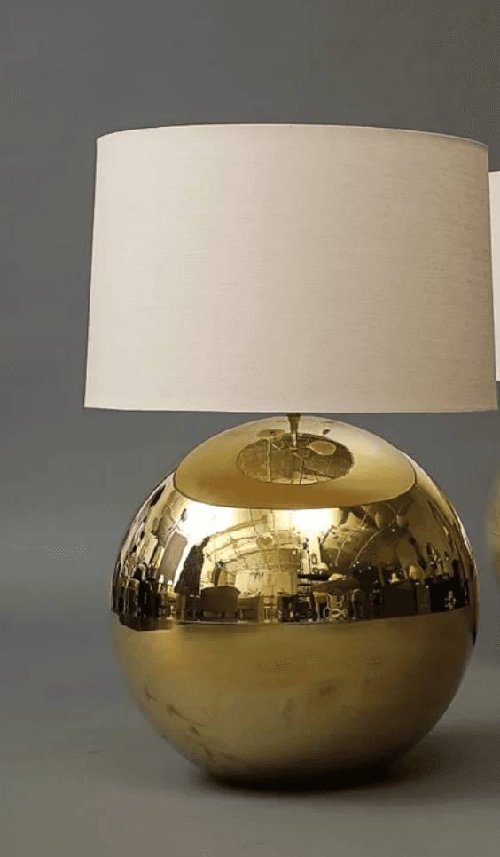 A table lamp featuring a spherical, highly polished brass-toned metal base and a plain, cylindrical… — 3D model