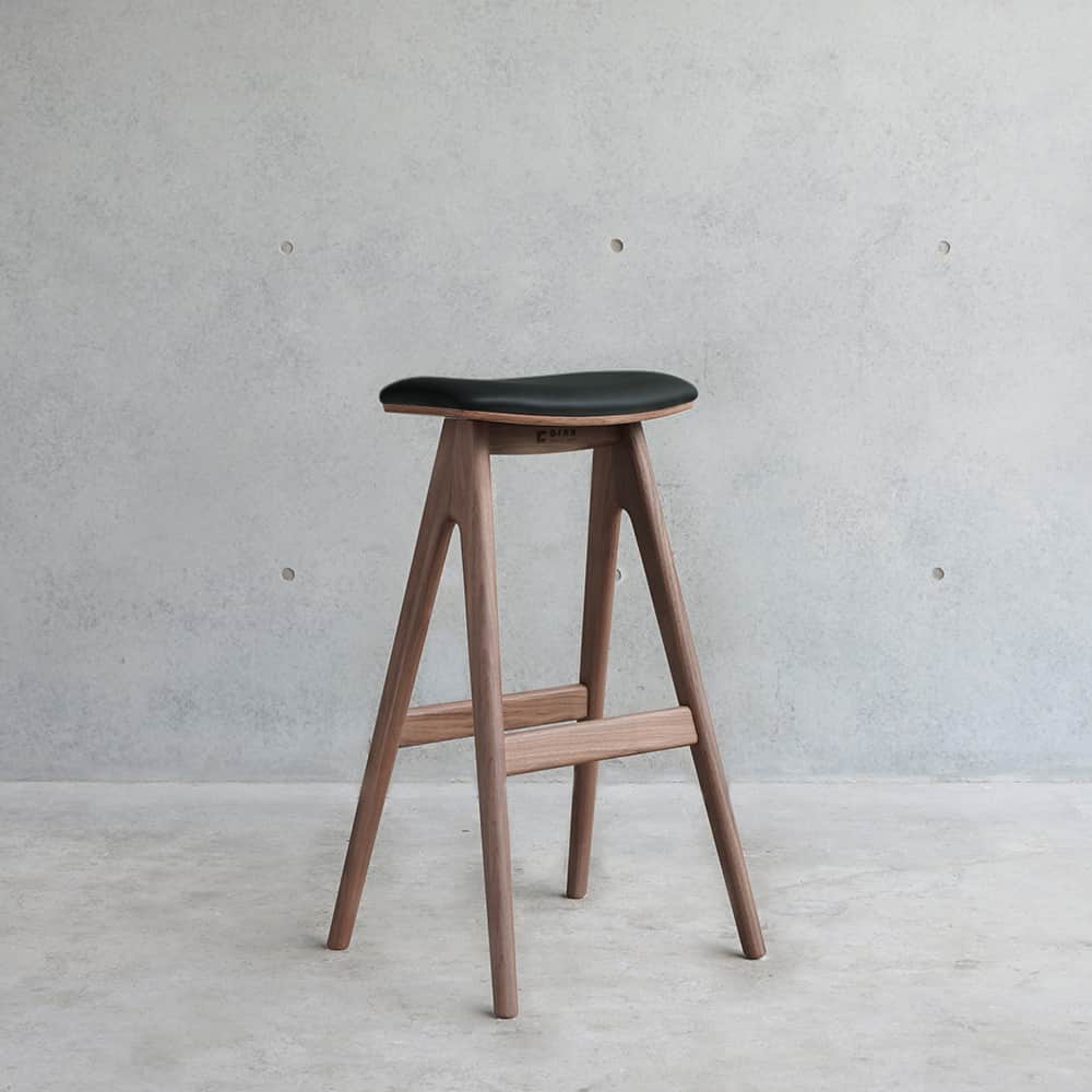 A modern bar stool with a saddle-shaped seat and a wooden frame. The seat is upholstered in black ma… — 3D model