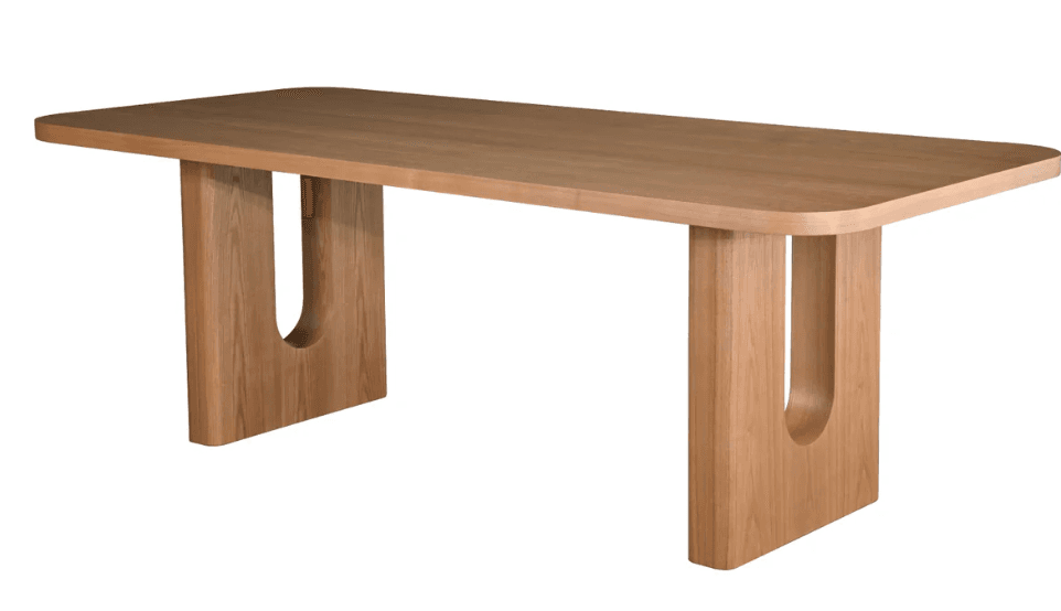 A modern dining table featuring a thick, rounded rectangular tabletop made from light-toned wood. The table is supported by two substantial pedestal legs, each characterized by a large, U-shaped cutout that provides visual interest and a sense of lightness. The overall aesthetic is minimalist and contemporary, with smooth surfaces and natural wood grain visible throughout. — 3D model