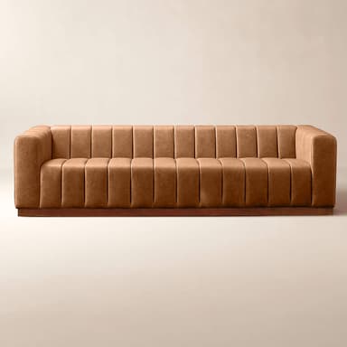 A long, channel-tufted sofa with a minimalist design. The sofa features a continuous padded back and armrest with vertical channels, upholstered in a tan leather-like material. It rests on a low, dark wood plinth base. The overall aesthetic is modern with a touch of art deco. — 3D model