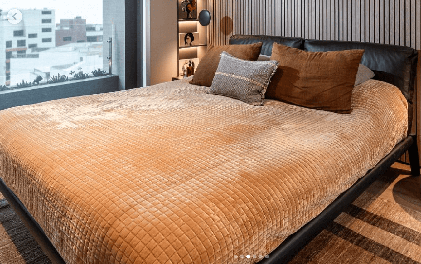 A modern platform bed featuring a dark, padded leather headboard with two distinct sections, supported by a minimalist, low-profile black frame and integrated tapered legs. The bed is adorned with a textured brown quilted coverlet and an assortment of brown and grey throw pillows. — 3D model