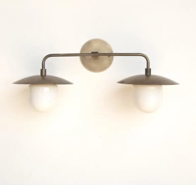 A wall-mounted lighting fixture featuring an aged brass finish, with two articulating arms extending horizontally from a circular backplate. Each arm supports a disc-shaped metal shade that directs light downwards onto an opaque white spherical diffuser. The design is minimalist and modern. — 3D model