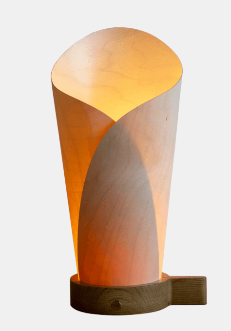 A modern table lamp featuring a sculptural shade crafted from bent plywood or wood veneer, which creates a warm, diffused light. The shade forms an organic, tapering shape, resting on a solid wood base. The design is minimalist and emphasizes natural materials. — 3D model