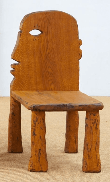 A distinctive carved wooden chair crafted from medium-brown oak, featuring an anthropomorphic backrest shaped like a human profile with a prominent eye cutout. The seat is a flat, solid square, supported by four chunky, irregularly shaped legs that contribute to its primitive, folk-art aesthetic. — 3D model
