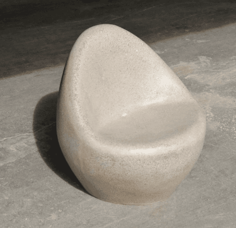 A single, smooth, stone-like chair with a rounded, organic shape. The chair features a scooped seat and a low backrest, resembling a pebble or a modern sculpture. Its design is minimalist and ergonomic, intended for single occupancy. The texture appears to be slightly granular or porous. — 3D model