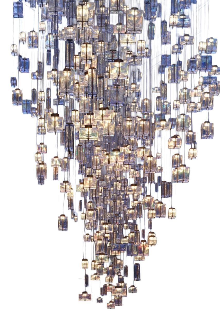 A grand, multi-tiered chandelier featuring numerous individual lighting elements suspended at varying heights to create a dramatic cascading effect. Each element consists of a rectangular block of textured or molded glass, appearing in shades of clear, blue, and amber, encasing an internal light source. The overall form tapers downwards, presenting a dynamic and luxurious illuminated sculpture. — 3D model