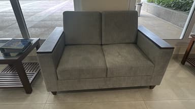 A modern two-seater loveseat with a light grey fabric upholstery and dark wood panels integrated into the top of the armrests. It features a clean, boxy silhouette with individual seat and back cushions. — 3D model