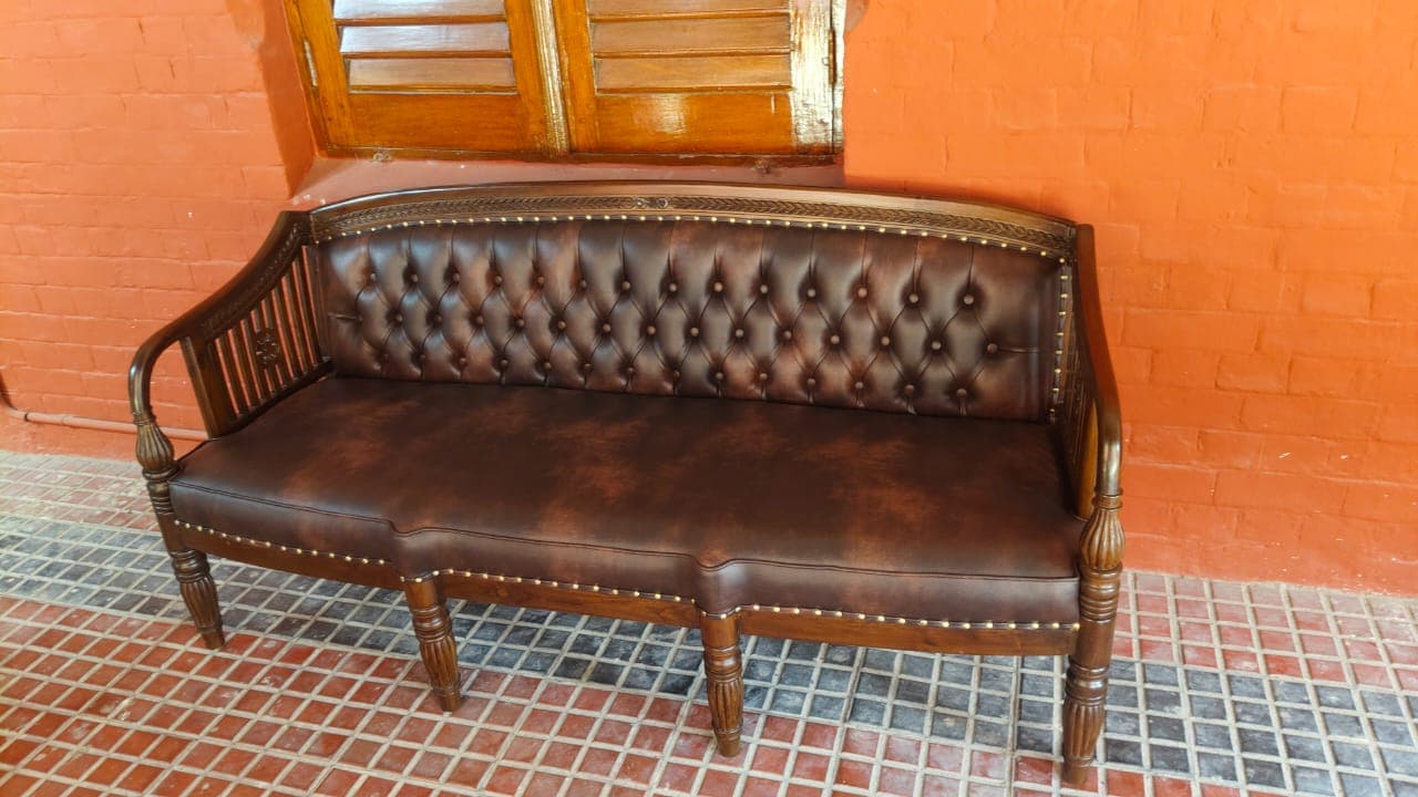 A traditional style three-seater sofa, featuring a dark brown wooden frame with intricate carvings and turned legs. The backrest and seat are upholstered in deep brown tufted leather, accented with brass nailhead trim. The armrests are an extension of the wooden frame, incorporating slatted vertical elements and decorative rosettes. — 3D model