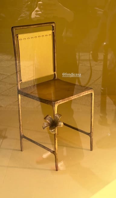 A decorative chair with a bronze-toned metal frame. The seat and backrest appear to be made of metal. The backrest features a panel of light tan leather or faux leather, attached with visible stitching. A prominent floral embellishment is attached to the front leg of the chair. The overall design suggests an artistic or decorative style. — 3D model