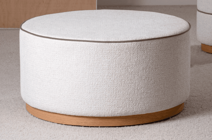 A contemporary round upholstered ottoman featuring a light-colored, textured fabric upper portion and a low, natural wood plinth base. A subtle dark brown piping detail is visible along the top edge where the seat meets the side. — 3D model