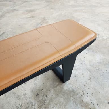 A long, rectangular bench featuring a padded, tan upholstered seat with stitched detailing, supported by a minimalist black metal frame with inverted V-shaped legs. — 3D model