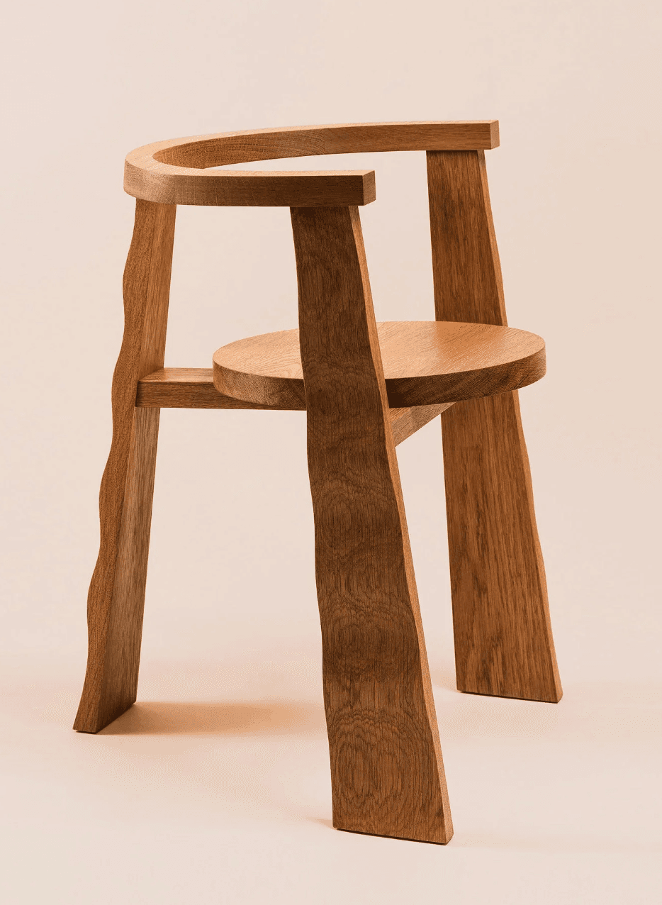 A contemporary solid wood chair featuring a distinctive sculptural form. It has a circular seat supported by three stout, irregularly shaped legs that are angled outwards. The backrest is formed by a curved piece of wood that wraps around, extending into a short armrest on one side, and is supported by a fourth leg that echoes the organic shape of the others. The natural wood grain is visible throughout. — 3D model