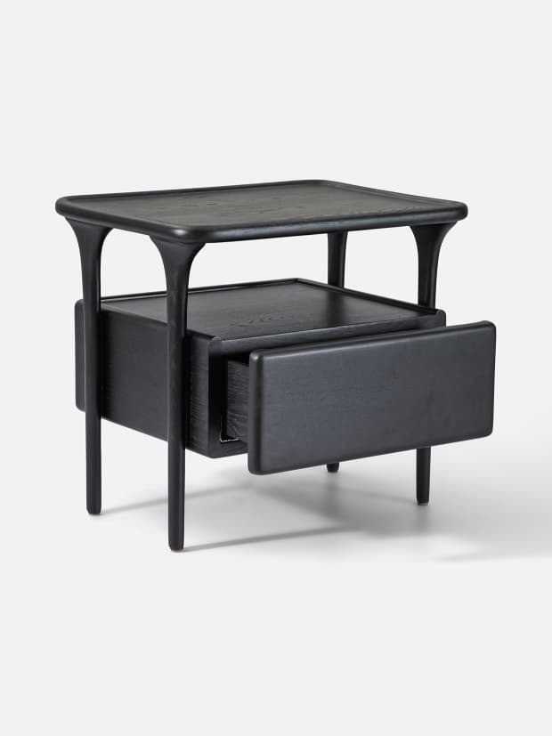 A modern nightstand crafted from black stained wood, featuring a rectangular top with subtly rounded edges, an open shelf below, and a single drawer. The drawer has a clean, handle-less front and appears to be cantilevered. The piece stands on four slender, tapered legs that extend upwards to support the open shelf and top surface. — 3D model