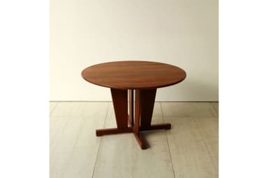 A round wooden table with a sturdy pedestal base, featuring a reddish-brown finish. The top is circular, and the base consists of a central column that flares out into an X-shaped foot structure on the floor, providing stable support. The design appears functional and clean-lined. — 3D model