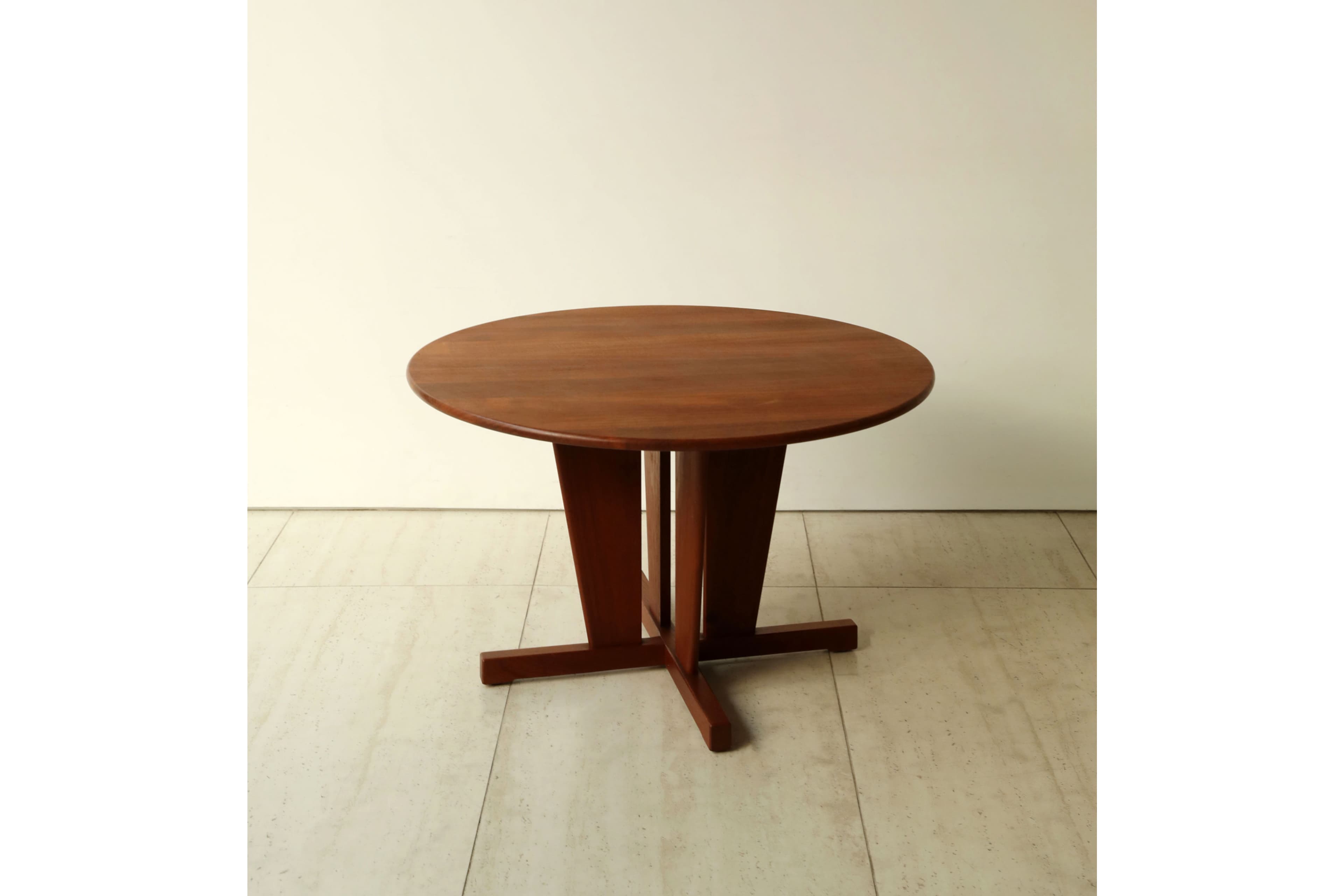 A round wooden table with a sturdy pedestal base, featuring a reddish-brown finish. The top is circular, and the base consists of a central column that flares out into an X-shaped foot structure on the floor, providing stable support. The design appears functional and clean-lined. — 3D model