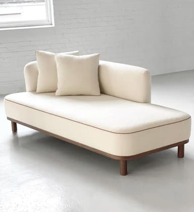 A contemporary cream-colored upholstered chaise lounge featuring a single raised armrest/backrest on the right side (when seated), a contrasting brown piping detail, and a dark wood base with short cylindrical legs. It includes two matching loose throw pillows. — 3D model