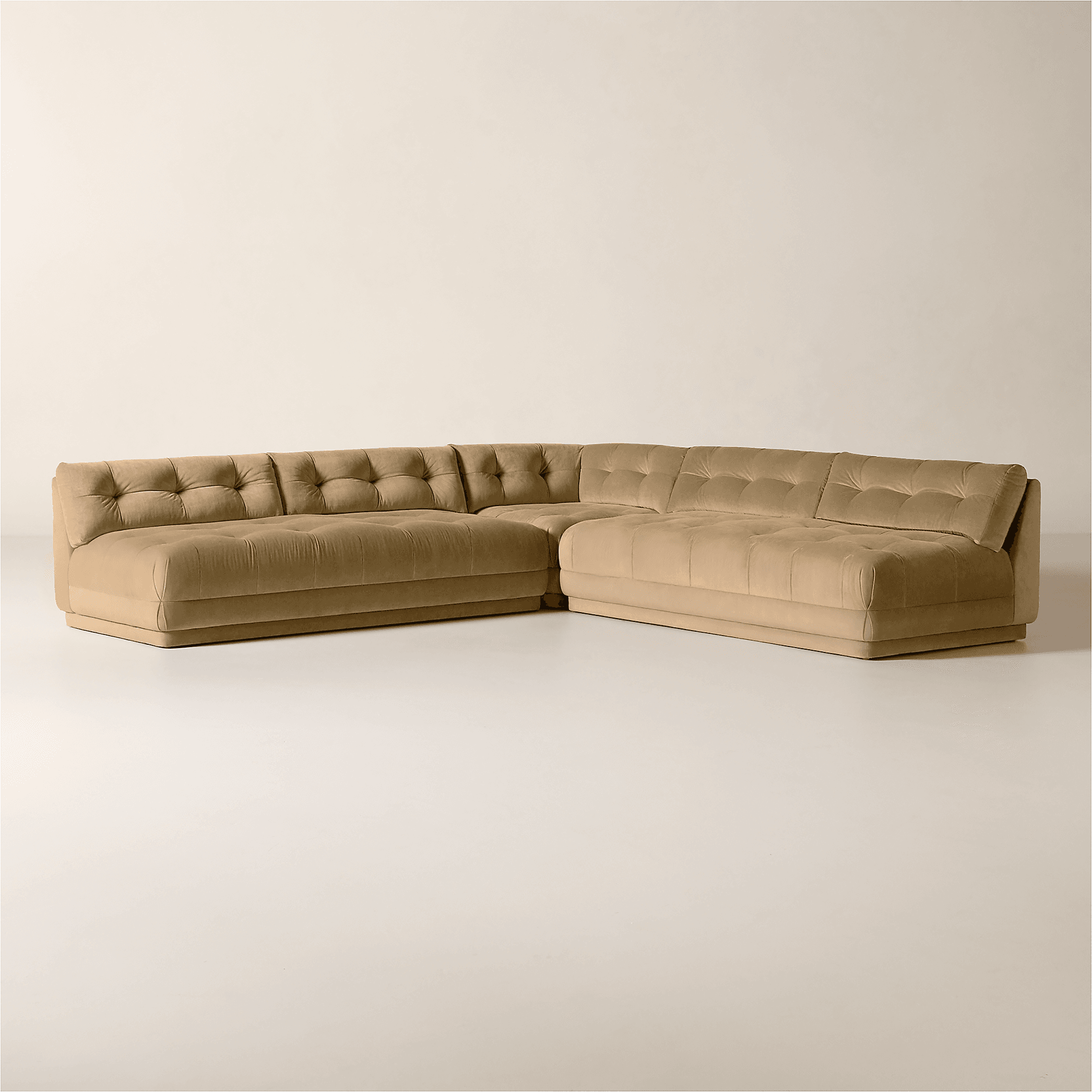 A tufted sectional sofa with a low profile and blocky silhouette. The sofa features a plush, velvet-like upholstery and deep button tufting on both the seat and back cushions, creating a series of geometric indentations. The arms are integrated into the frame, with one arm angled slightly outwards on the right side. The base is solid and appears to be a continuous platform. The color is a neutral, warm beige. — 3D model