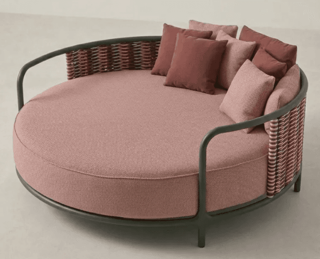 A contemporary outdoor circular daybed featuring a dark green metal frame, a woven backrest in variegated tones of pink and brown, and a large round cushion with multiple throw pillows in shades of pink and rose. The base cushion and some pillows have a textured fabric finish, while others are solid. — 3D model