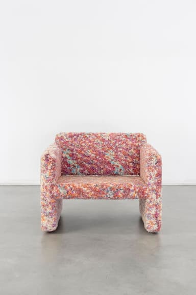 A contemporary, low-profile armchair featuring a distinctive multicolored, highly textured upholstery. The chair has a blocky, sculptural silhouette with integrated arms and backrest that flow seamlessly into the seat and base, giving it a continuous, monolithic appearance. The fabric presents a vibrant, speckled pattern composed of various hues including pink, red, orange, blue, purple, and yellow, creating a playful and eye-catching aesthetic. The base is also fully upholstered, eliminating visible legs. — 3D model