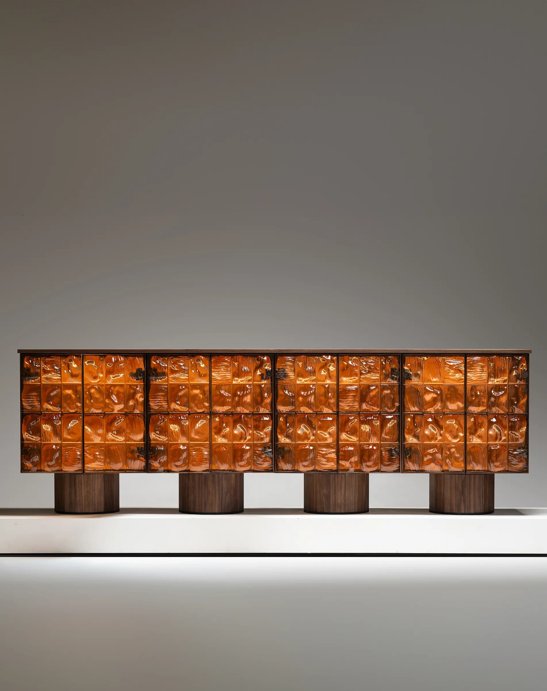 A long, low cabinet with a dark wood frame. The front of the cabinet is composed of multiple rows and columns of textured, amber-colored glass panels, which appear to be illuminated from within. The cabinet is supported by four cylindrical wooden legs. — 3D model