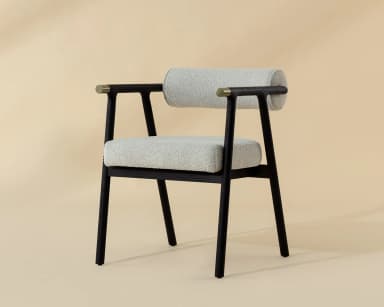 A contemporary armchair featuring a dark, possibly black-stained, wooden frame with clean lines. The seat and cylindrical backrest are upholstered in a light, textured fabric, resembling bouclé or a similar weave. The armrests are an extension of the frame, culminating in subtle brass-finished accents on the outer edges. The chair's design combines minimalist aesthetics with comfortable padding, suitable for various modern interiors. — 3D model