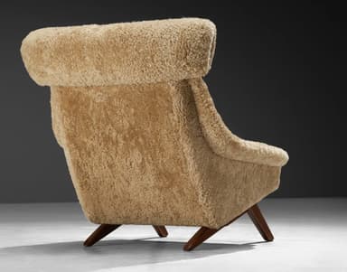 A high-backed armchair with a distinctive sloped silhouette. The chair is upholstered entirely in a textured, light brown sheepskin or shearling material, giving it a plush and organic appearance. The seat and back cushions are integrated into the overall form. The base consists of dark wood legs with a visible grain, angled outwards for stability. The headrest area is pronounced and rolls over the top of the backrest. — 3D model