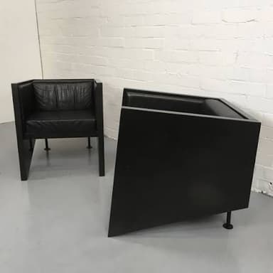 A pair of modern armchairs with a distinctive geometric design, featuring angular dark-stained wood or laminate frames and black leather seat and back cushions. The chairs present a minimalist and architectural aesthetic. — 3D model