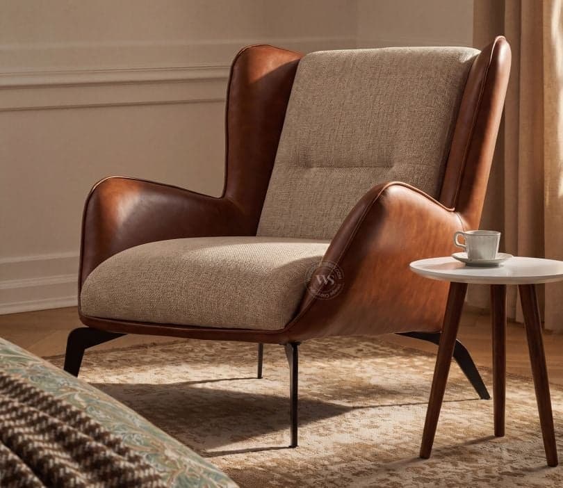 A modern lounge chair featuring a distinctive two-tone upholstery design. The outer shell and armrests are covered in smooth brown leather, while the inner seat, backrest, and side panels are upholstered in a textured light beige fabric. The chair has a comfortable, slightly reclined back with a single button tuft on the lower back. It stands on slender, splayed dark metal legs. — 3D model