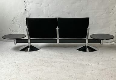 A two-seater sofa with attached side tables. The sofa features black upholstered seats and backrests with a ribbed texture on the backrests. The frame appears to be metal, possibly chrome or stainless steel, with a dark finish on the main horizontal support. Each seat has a cantilevered base with a circular, flared chrome pedestal. Attached to the horizontal frame on either side are circular, dark-colored side tables with a glossy finish, supported by metal brackets. — 3D model