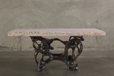 A console table with a rectangular, light pinkish-beige stone top and an intricately sculpted, dark bronze base. The base features an organic, abstract design with interconnected hollow forms and flowing lines, resembling bones or skeletal structures. The stone top has a rough-hewn edge and a textured surface with visible inclusions. — 3D model