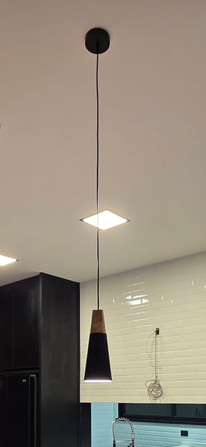 A minimalist pendant light featuring a cylindrical black canopy connected to a thin black suspension cable. The main light fixture consists of a natural wood element that tapers into a matte black metal cone-shaped shade. Above the shade, a small, flat, square-shaped diffuser with rounded corners provides additional downward illumination, indicating an integrated LED design. The light fixture has a clean, modern aesthetic with a combination of natural and industrial materials. — 3D model