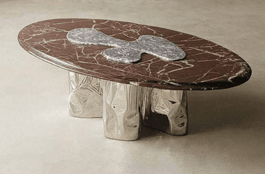 An oval-shaped coffee table featuring a polished brown marble top with prominent white veining. An abstract, irregular-shaped inlay of lighter grey stone is centered on the tabletop. The table is supported by a sculptural, three-pronged metallic base with an organic, uneven, and reflective surface finish, likely polished chrome or nickel, giving it a fluid appearance. — 3D model