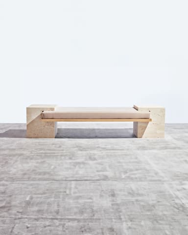 A contemporary bench featuring two rectangular, textured travertine stone bases supporting a horizontal upholstered seat. A thin, metallic frame, possibly brass or gold-toned, is visible beneath the cushion, connecting the two stone supports. The travertine exhibits natural pitting and veining characteristic of the material. The seat cushion is a neutral, light-colored fabric. — 3D model