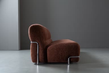 A compact, armless loveseat upholstered in a textured, dark reddish-brown fabric, featuring a minimalist design with a chrome-finished metal bar accent along the base. — 3D model