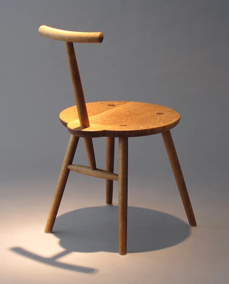 A wooden chair with a round seat and a single backrest support. The chair has four legs, with two legs at the front and two at the back. There is a horizontal wooden brace connecting the front and back legs on each side. The backrest is a curved piece of wood supported by a vertical rod extending from the seat. The seat is slightly dished and shows visible wood grain. — 3D model