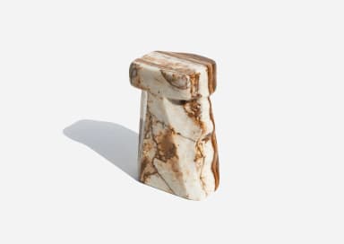 A small, monolithic piece of furniture carved from a single block of variegated marble, featuring a flat, octagonal-like top surface supported by a thick, sculpted base with a distinct 'T' or inverted 'L' profile and subtly faceted sides. The stone displays natural white, cream, and beige tones with prominent brown, rust, and grey veining. — 3D model