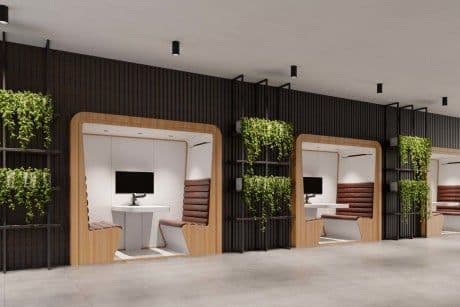 An isolated, isometric view of a modern acoustic office privacy pod designed for individual work or small meetings. The pod features a light wood exterior with rounded edges, a white laminate roof, and an interior fitted with brown horizontally-tufted upholstered seating on opposing sides, separated by a white integrated tabletop. A small monitor is mounted on one interior wall. One exterior side is equipped with a black metal grid structure holding planters with cascading green foliage, providing a biophilic design element. The base and interior walls are white, contrasting with the wood and upholstery. — 3D model