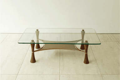 A modern coffee table featuring a rectangular clear glass top with softly rounded corners. The base consists of four sculptural, inverted-hourglass-shaped wooden legs, likely walnut, accented with brass rings. These legs support a brass frame that cradles a beige or taupe canvas sling, forming a secondary storage or display surface beneath the glass. The overall design suggests a blend of organic forms and metallic accents. — 3D model