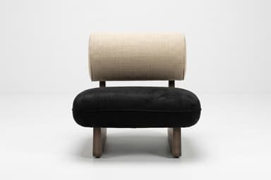 A modern, low-profile lounge chair featuring a distinctive solid wood base. The base is dark-toned wood, shaped with a geometric, wedge-like profile. It supports a plush, rectangular black seat cushion, likely upholstered in velvet or microsuede. The backrest is a cylindrical bolster cushion, upholstered in a textured light beige fabric, resting against the angled wooden support structure. — 3D model