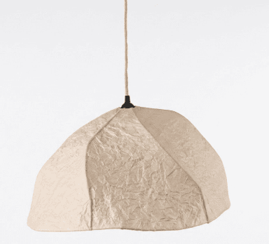 A contemporary pendant light featuring an irregularly shaped, organic dome-like shade made from highly textured, crumpled light beige or cream fabric. The shade is suspended by a visible braided rope cord, connected to a small black ceiling mount or fixture at the top. The overall design emphasizes natural textures and a soft, sculptural form. — 3D model