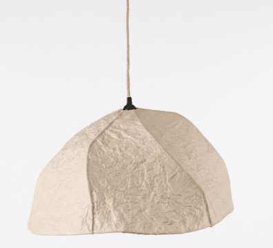 A contemporary pendant light featuring an irregularly shaped, organic dome-like shade made from highly textured, crumpled light beige or cream fabric. The shade is suspended by a visible braided rope cord, connected to a small black ceiling mount or fixture at the top. The overall design emphasizes natural textures and a soft, sculptural form. — 3D model
