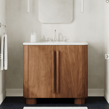 A modern bathroom vanity featuring a solid wood or wood veneer cabinet with a white ceramic integrated sink and countertop. The cabinet has rounded edges and two doors with recessed vertical bar handles. It stands on four short, cylindrical block feet. A chrome faucet and a white soap dispenser are visible on the countertop. — 3D model