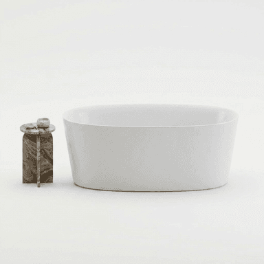 A freestanding, oval-shaped bathtub with a smooth, matte white exterior and interior. The tub has a solid, seamless construction with a gently sloping rim. It is positioned next to a small, circular side table with a multi-colored, veined stone top and a cross-shaped base. Two small white candles and a dish are placed on the side table. — 3D model