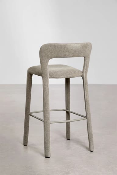 A modern bar stool with a slightly curved backrest and a seat upholstered in a textured beige fabric. The stool has four tapered legs that extend upwards to form the seat and backrest structure. Metal footrests are integrated between the legs. — 3D model