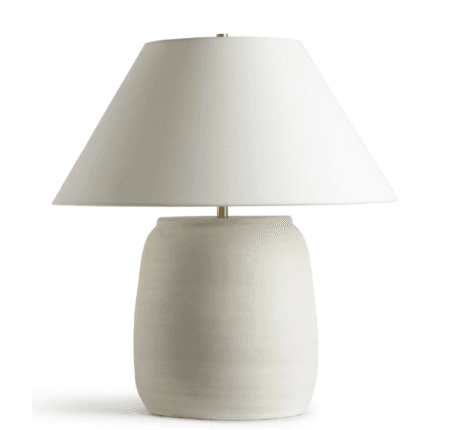 A modern table lamp featuring an off-white, textured ceramic base with a slightly barrel-shaped form, topped with a matching off-white, conical fabric shade. Small brass accents are visible at the neck and finial, complementing the minimalist design. — 3D model