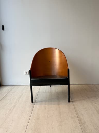 A striking mid-century modern lounge chair featuring a gracefully curved, light-brown plywood shell that forms both the backrest and integrated armrests. The chair is supported by a minimalist black metal frame with slender legs. A dark, possibly black or dark brown, upholstered cushion rests on the seat, contrasting with the warm wood tones. — 3D model