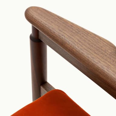 A contemporary armchair featuring a dark wood frame and rust-colored upholstered seat and backrest. The backrest is rectangular with cut-outs to accommodate the rounded wooden armrests, which extend from the rear support posts. The legs are cylindrical and straight, connected by stretcher bars beneath the seat. The seat cushion is subtly tufted. — 3D model