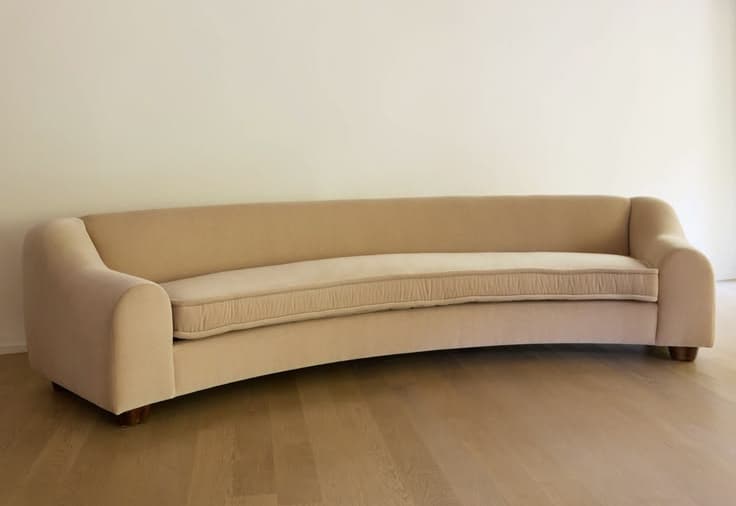 A modern curved sofa featuring a beige upholstered fabric, low profile, and simple block feet. The design includes fixed back and seat cushions, with gently rolled arms that flow into the curved backrest. — 3D model
